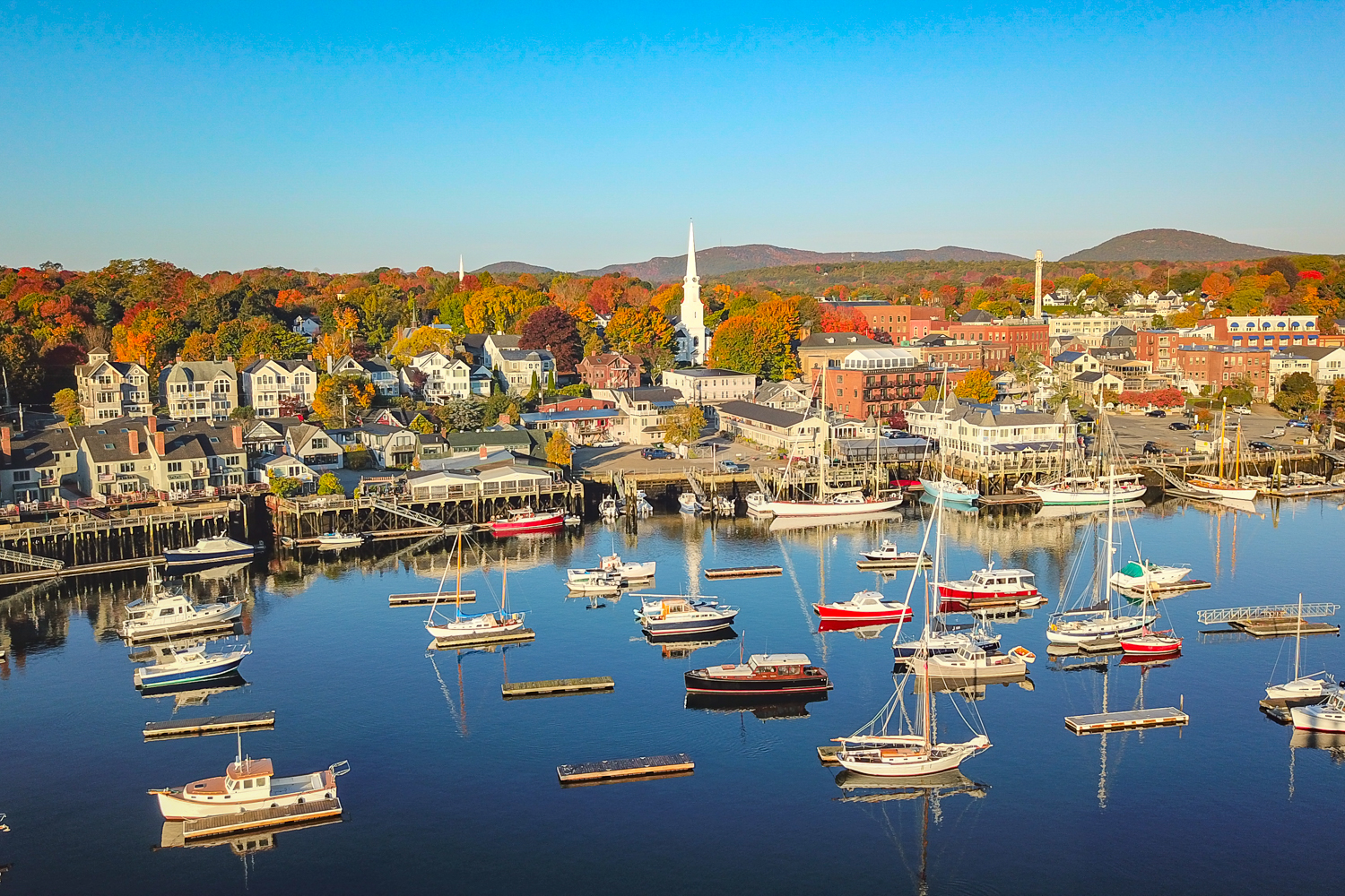 Aerial view of Camden Harbor, Maine with sailboats, waterfront homes, fall foliage, and downtown village—coastal Maine real estate and buying and selling homes in Maine.
