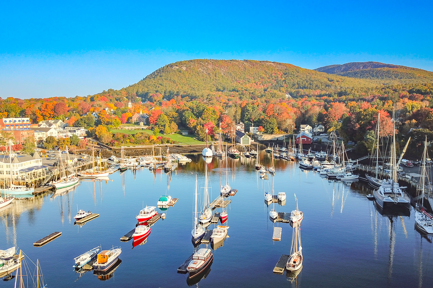 Aerial view of Camden Harbor, Maine with sailboats, waterfront homes, fall foliage, and downtown village—coastal Maine real estate and buying and selling homes in Maine.