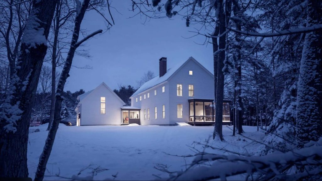 White Maine farmhouse surrounded by snow at dusk with warm interior lights glowing through the windows.