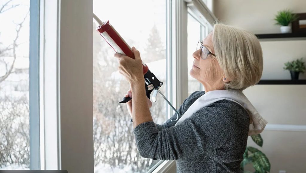 Homeowner applying caulk around a window frame to prevent winter drafts and improve energy efficiency.
