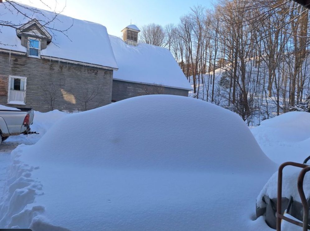 Vehicle nearly buried under deep snow beside a Maine barn after a heavy winter storm.