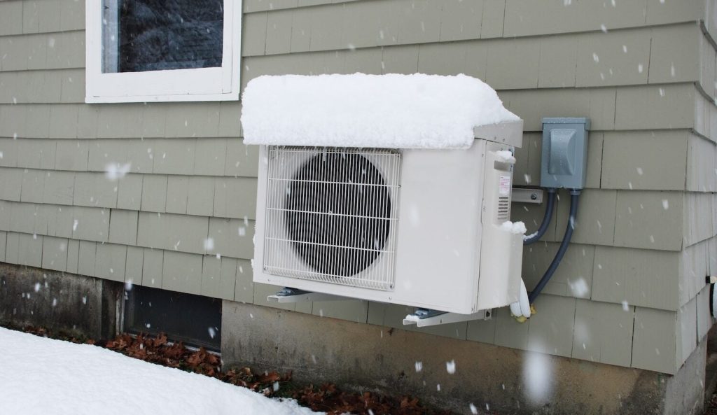 Cold-climate heat pump mounted on the exterior of a Maine home during active snowfall.