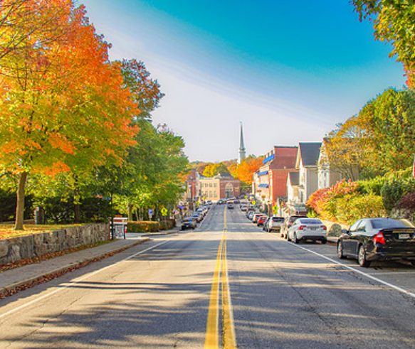 Downtown Camden Maine in the fall with colorful trees, parked cars, and a view down Main Street toward the harbor.