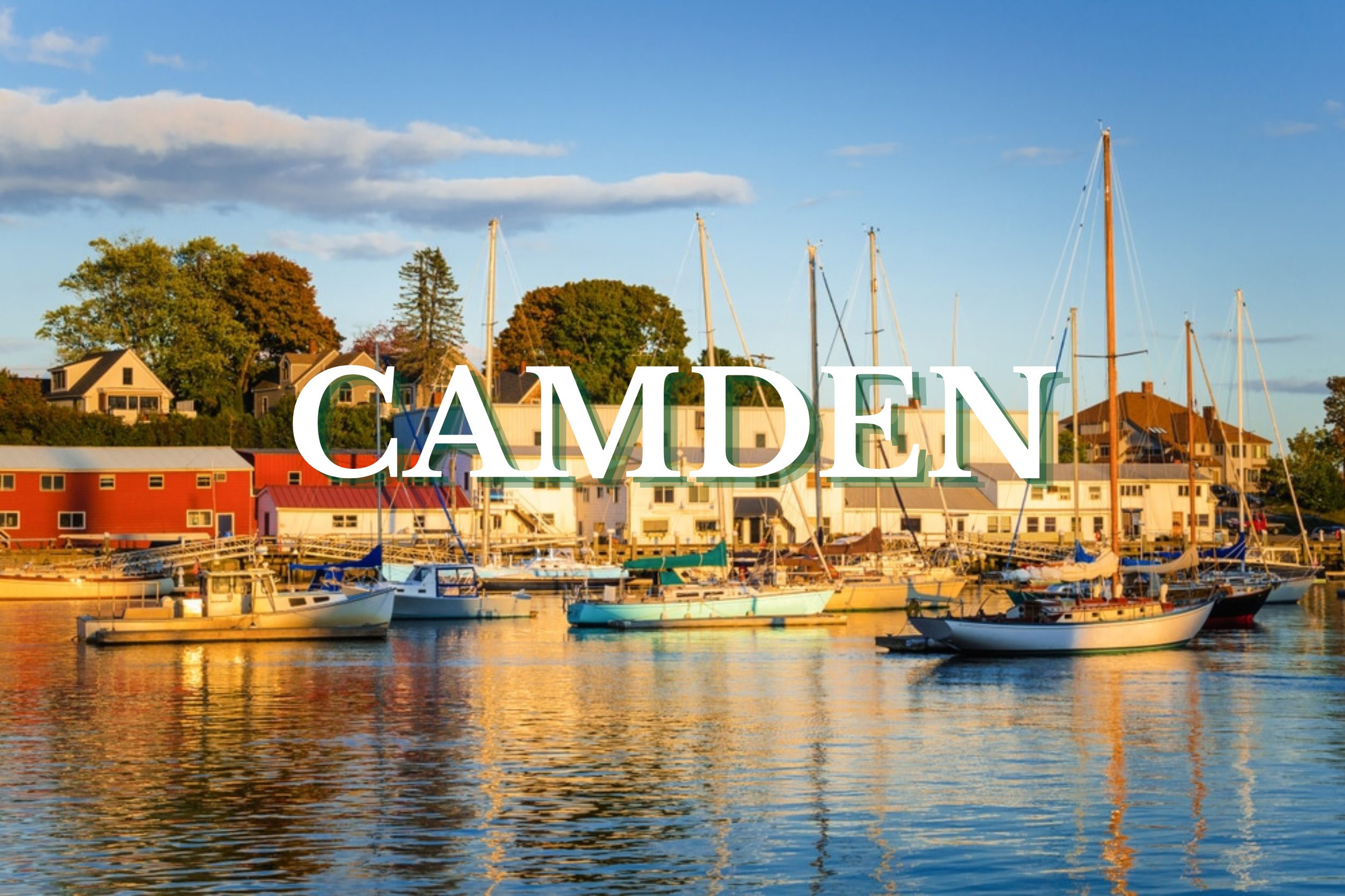Sailboats docked in Camden Harbor with waterfront buildings and blue skies in Camden, Maine