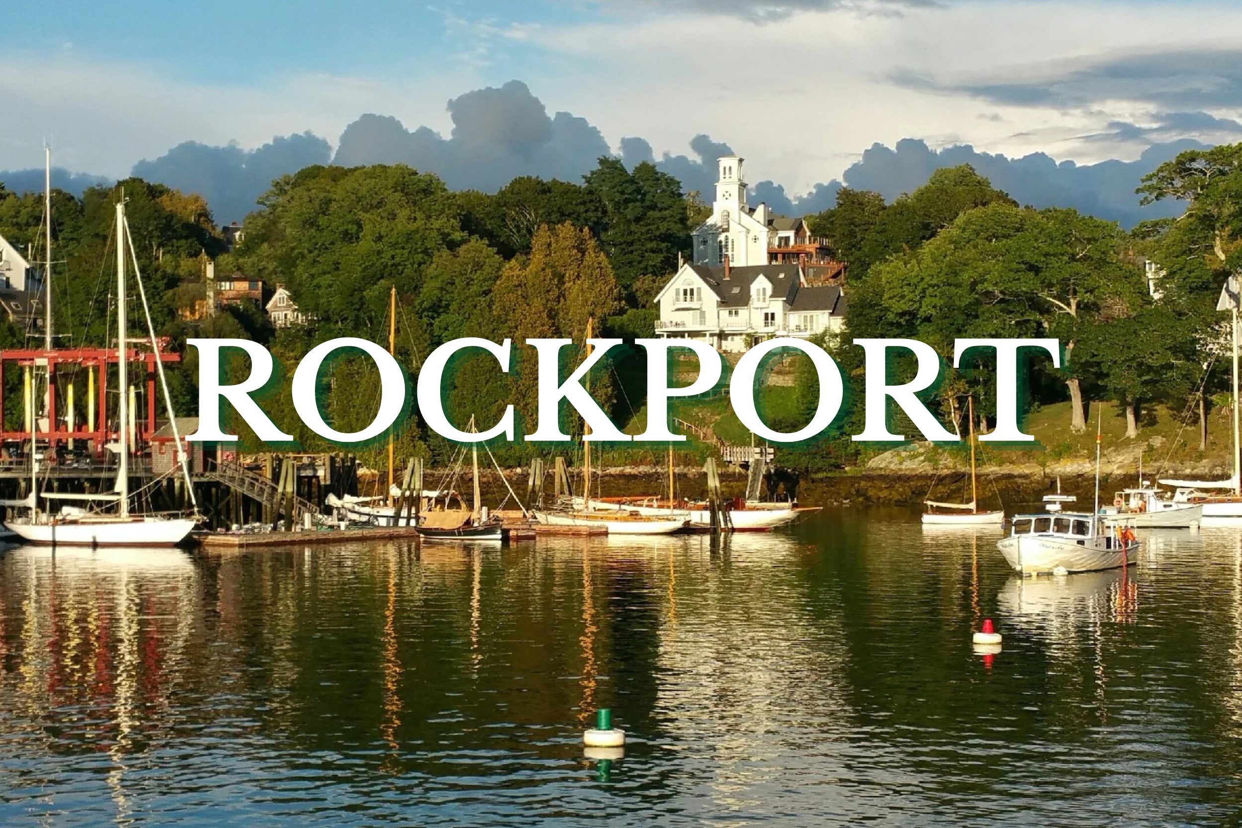 Boats docked along the peaceful harbor waterfront in Rockport, Maine with coastal homes and trees in the background