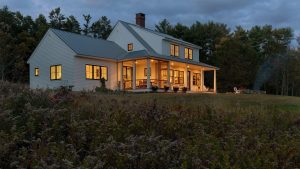 White Maine farmhouse with a covered porch glowing at dusk and surrounded by trees